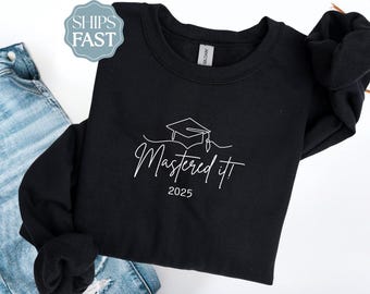 May include: Black sweatshirt with the text "Mastered It!" and the year 2025 in white script. A graduation cap graphic is above the text. The sweatshirt is folded, with a pair of blue jeans in the background.
