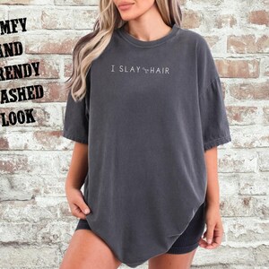May include: A dark gray oversized t-shirt with the text "I SLAY HAIR" printed on the front. The shirt has a relaxed fit and a washed look.