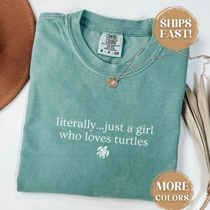 May include: A seafoam green t-shirt with the text "literally...just a girl who loves turtles" in white. A small turtle graphic is below the text. A silver necklace with a pendant is on the shirt.