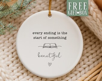 May include: A white ceramic ornament with the text "every ending is the start of something beautiful" and a small heart. The ornament has a gold string for hanging and is set against a cozy, neutral-toned background.