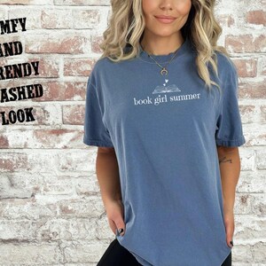 May include: A blue jean-colored t-shirt with the text "book girl summer" in white. The shirt has a relaxed fit and is worn by a person. The words "Comfy and Trendy Washed Look" are written on the left side of the image.