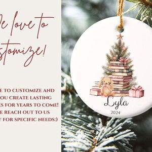 May include: A white ceramic Christmas ornament with a watercolor illustration of a Christmas tree decorated with books and a small bunny. The ornament is personalized with the name "Lyla" and the year "2024".