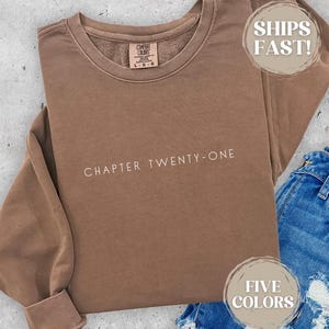 May include: A brown sweatshirt with the text "CHAPTER TWENTY-ONE" in white. The sweatshirt is folded, with a pair of blue jeans visible in the bottom right corner. The sweatshirt has a crew neck and long sleeves.