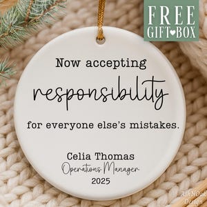 May include: A white ceramic ornament with the text "Now accepting responsibility for everyone else's mistakes." The ornament includes the name "Celia Thomas", "Operations Manager", and the year "2025". A gold string is attached for hanging.