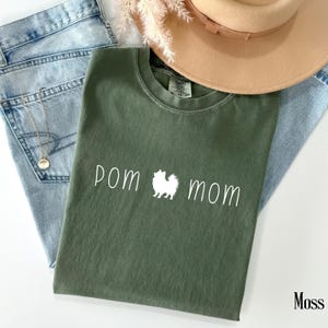 May include: A moss green t-shirt with the words "pom mom" in white script, with a white Pomeranian dog silhouette between the words. The shirt is laid on top of light blue jeans, a gold necklace, and a tan hat.