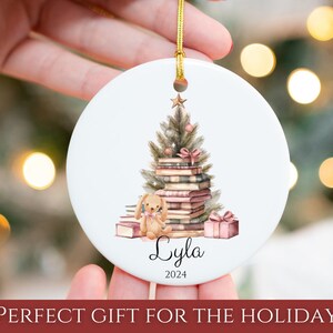 May include: A white ceramic Christmas ornament with a watercolor illustration of a Christmas tree made of stacked books. A small, brown teddy bear sits at the base of the tree. The ornament is personalized with the name "Lyla" and the year "2024".
