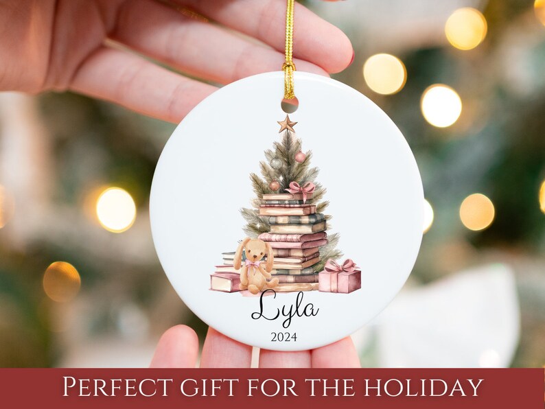 May include: A white ceramic Christmas ornament with a watercolor illustration of a Christmas tree made of stacked books. A small, brown teddy bear sits at the base of the tree. The ornament is personalized with the name "Lyla" and the year "2024".