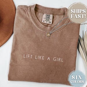 May include: A brown t-shirt with the text "LIFT LIKE A GIRL" printed in white. The shirt is made of a soft, comfortable material.