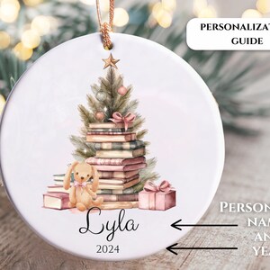 May include: A white ceramic ornament with a watercolor illustration of a Christmas tree made of books, a small bunny, and a gift box. The text "Lyla 2024" is written below the illustration.