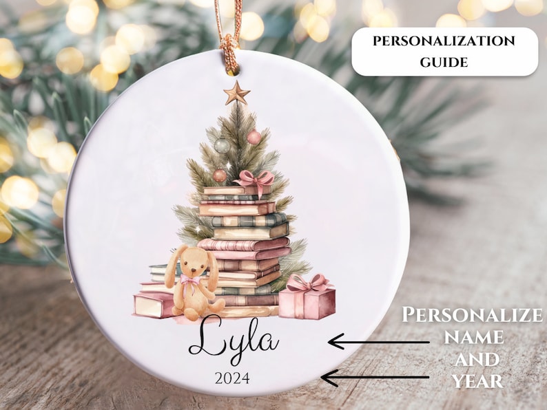 May include: A white ceramic ornament with a watercolor illustration of a Christmas tree made of books, a small bunny, and a gift box. The text "Lyla 2024" is written below the illustration.