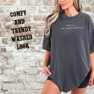 May include: Dark gray t-shirt with the text "hold on..... my ragdoll needs me." The shirt has a washed look and a relaxed fit. The image also includes the text "COMFY AND TRENDY WASHED LOOK."