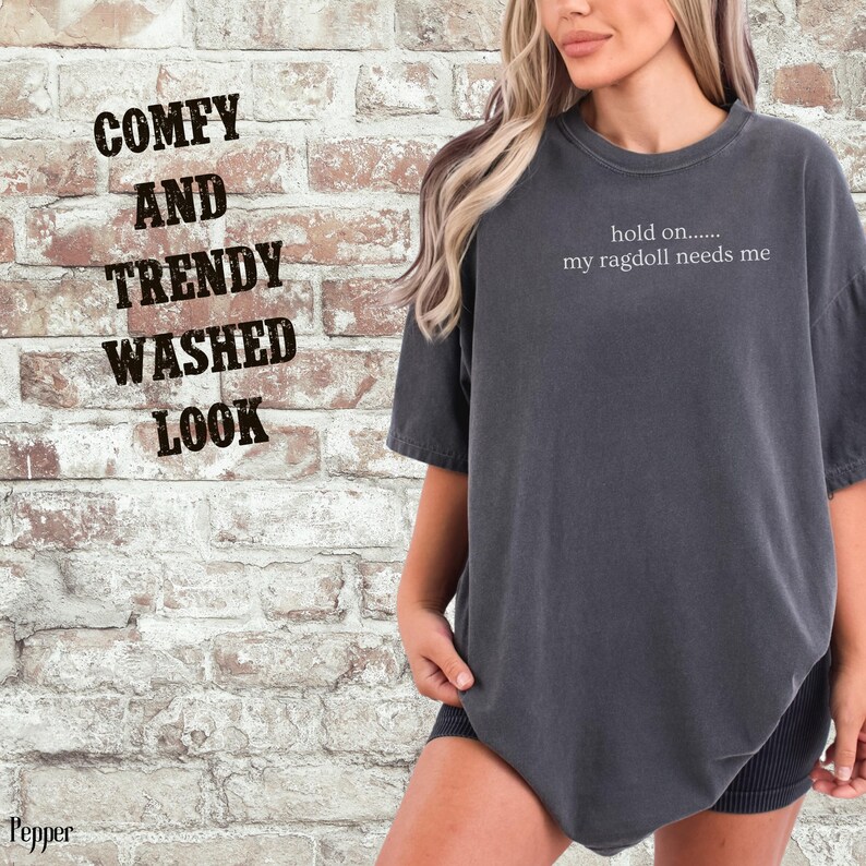 May include: Dark gray t-shirt with the text "hold on..... my ragdoll needs me." The shirt has a washed look and a relaxed fit. The image also includes the text "COMFY AND TRENDY WASHED LOOK."