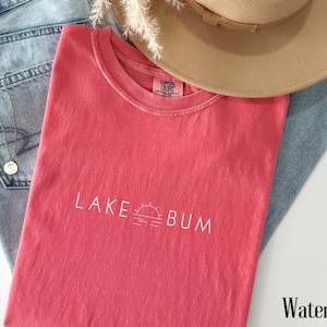 K&ouml;nnte beinhalten: Ein wasserfarbenes T-Shirt mit dem wei&szlig;en Aufdruck "LAKE BUM" und einer Sonne- und Wasser-Grafik. Das Shirt liegt auf einer blauen Jeans, mit einem beigen Hut und einem dekorativen Zweig.