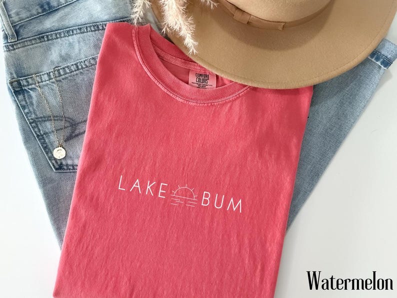 K&ouml;nnte beinhalten: Ein wasserfarbenes T-Shirt mit dem wei&szlig;en Aufdruck "LAKE BUM" und einer Sonne- und Wasser-Grafik. Das Shirt liegt auf einer blauen Jeans, mit einem beigen Hut und einem dekorativen Zweig.