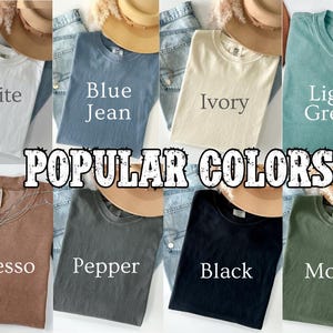 May include: A collection of eight t-shirts in various colors, including white, blue jean, ivory, light green, espresso, pepper, black, and moss. Each shirt is folded and displayed with a hat and denim jeans. The text "POPULAR COLORS" is overlaid.