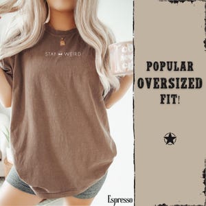 以下が含まれることがあります： 前面に「STAY WEIRD」とプリントされたエスプレッソカラーのオーバーサイズTシャツ。 ゆったりとしたフィット感と半袖です。 画像にはマグカップと「POPULAR OVERSIZED FIT！」の文字も表示されています。