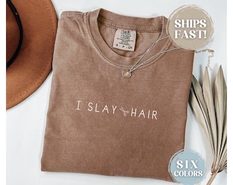 May include: A brown t-shirt with the text "I SLAY HAIR" in white lettering. The shirt has a distressed look and a silver chain necklace with a pendant.