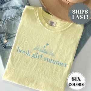 May include: A light yellow t-shirt with the text "book girl summer" in light blue, along with an open book graphic and two small hearts. The shirt is laid on top of blue jeans and a tan hat.