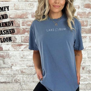 K&ouml;nnte beinhalten: Blaues Jeans-T-Shirt mit dem wei&szlig;en Text "LAKE BUM". Das Shirt hat einen Used-Look und einen bequemen, trendigen Stil. Das Bild enth&auml;lt auch den Text "COMFY AND TRENDY WASHED LOOK" und "Blue Jean".