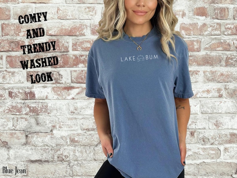 K&ouml;nnte beinhalten: Blaues Jeans-T-Shirt mit dem wei&szlig;en Text "LAKE BUM". Das Shirt hat einen Used-Look und einen bequemen, trendigen Stil. Das Bild enth&auml;lt auch den Text "COMFY AND TRENDY WASHED LOOK" und "Blue Jean".