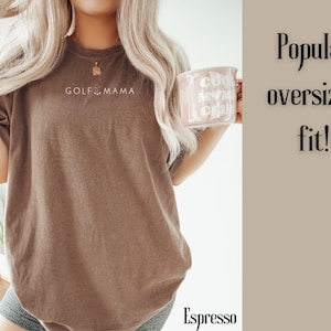 K&ouml;nnte beinhalten: Ein braunes Oversize-T-Shirt mit dem Aufdruck "GOLF MAMA" auf der Vorderseite. Das Shirt wird von einer Person getragen, die eine Kaffeetasse h&auml;lt.