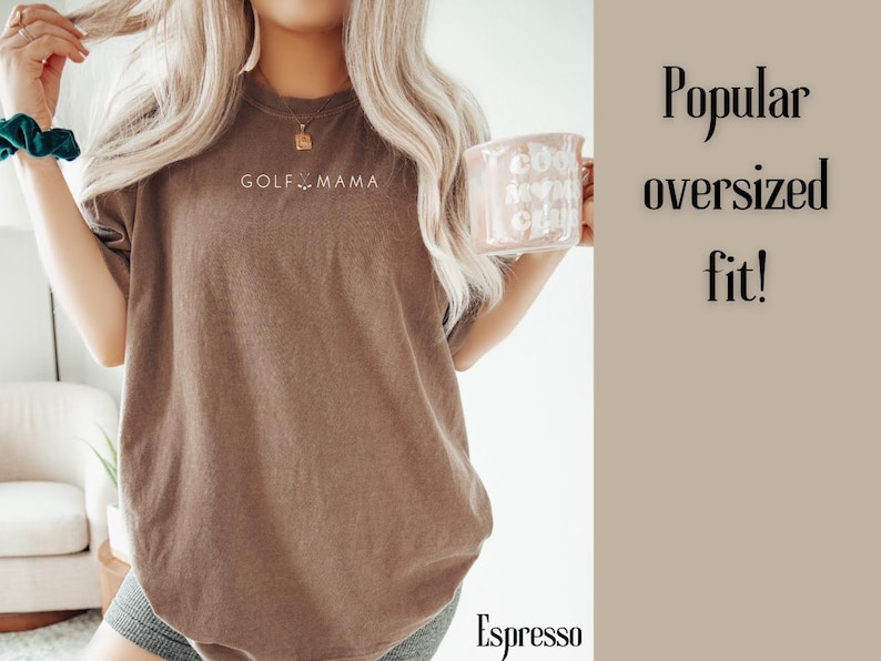 K&ouml;nnte beinhalten: Ein braunes Oversize-T-Shirt mit dem Aufdruck "GOLF MAMA" auf der Vorderseite. Das Shirt wird von einer Person getragen, die eine Kaffeetasse h&auml;lt.