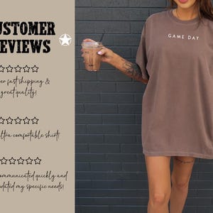 May include: A brown oversized t-shirt with the text "GAME DAY" printed on the front. The shirt is being worn by a person with tattoos on their arm. The background is a brick wall.