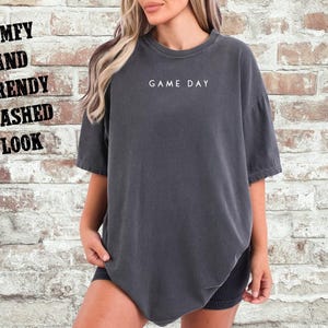 May include: A dark gray oversized t-shirt with the text "GAME DAY" printed in white on the front. The shirt has a relaxed fit and a washed look.