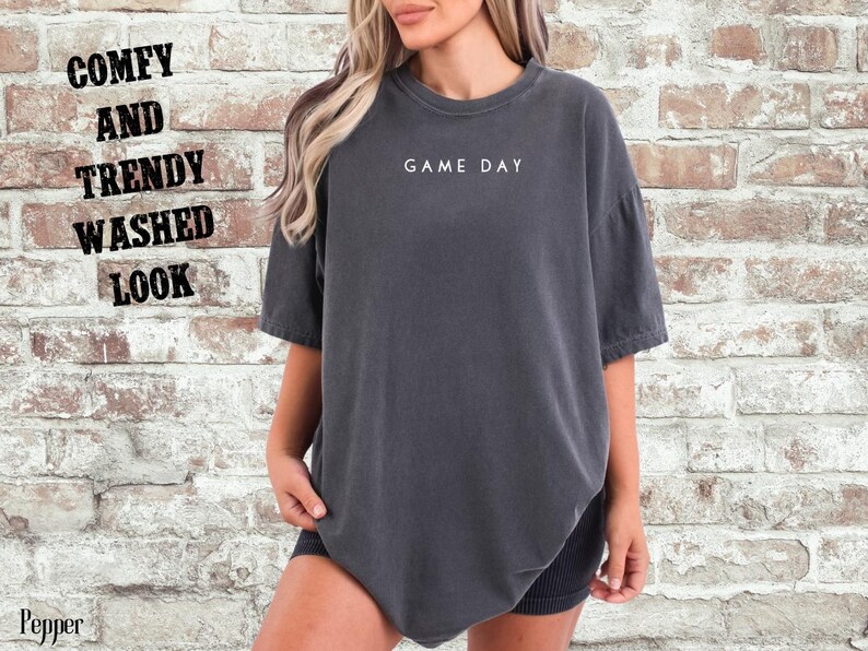 May include: A dark gray oversized t-shirt with the text "GAME DAY" printed in white on the front. The shirt has a relaxed fit and a washed look.
