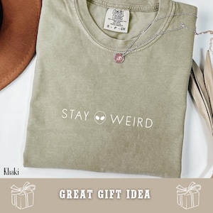 以下が含まれることがあります： カーキ色のTシャツ。白で「STAY WEIRD」の文字と宇宙人のグラフィックがプリントされています。小さなペンダントが付いたシルバーのネックレスがシャツの上にあります。シャツにはComfort Colorsのタグが付いています。「GREAT GIFT IDEA」の文字も表示されています。