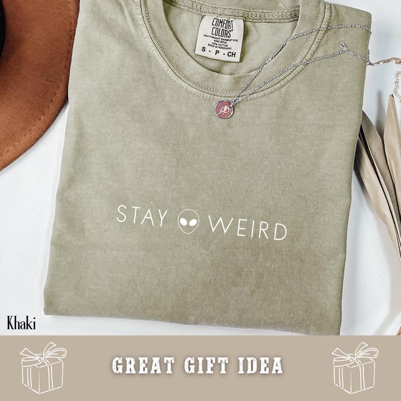 以下が含まれることがあります： カーキ色のTシャツ。白で「STAY WEIRD」の文字と宇宙人のグラフィックがプリントされています。小さなペンダントが付いたシルバーのネックレスがシャツの上にあります。シャツにはComfort Colorsのタグが付いています。「GREAT GIFT IDEA」の文字も表示されています。