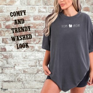 May include: A dark gray t-shirt with the words "pom mom" and a Pomeranian dog graphic. The shirt has a washed look and the words "Comfy and Trendy Washed Look" are printed on the brick wall background.
