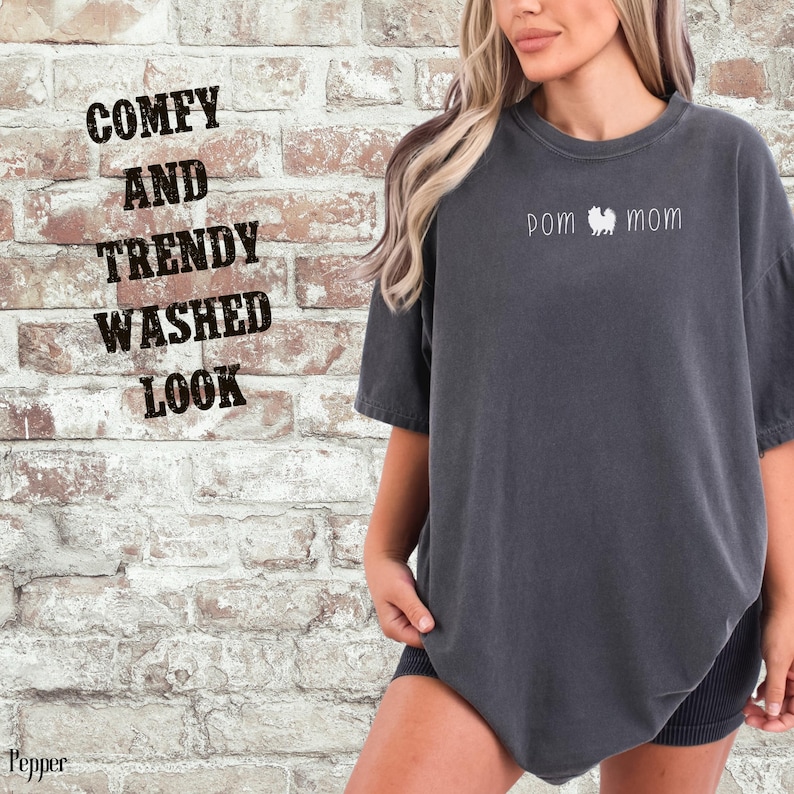 May include: A dark gray t-shirt with the words "pom mom" and a Pomeranian dog graphic. The shirt has a washed look and the words "Comfy and Trendy Washed Look" are printed on the brick wall background.