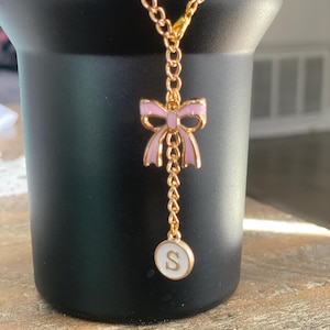 May include: A gold chain necklace with a pink bow charm and a white circle charm with the letter 'S' on it.