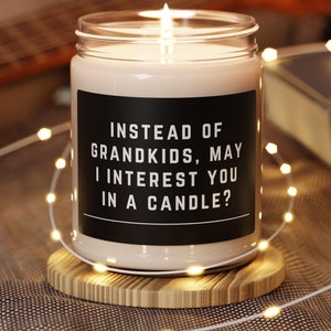 May include: A white candle with a black label that reads "Instead of grandkids, may I interest you in a candle?" The candle is sitting on a wooden coaster and surrounded by white string lights.