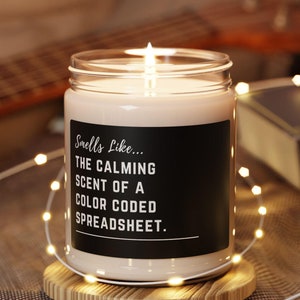 Op de afbeelding: Een witte kaars in een glazen pot met een zwart label waarop staat "Smells Like... THE CALMING SCENT OF A COLOR CODED SPREADSHEET."