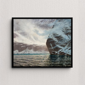 May include: A framed landscape print depicts a coastal scene with a large, snow-covered rock formation on the right. The sky is filled with dramatic, swirling clouds, and the water is a deep blue-green. The artwork is framed in a black border.