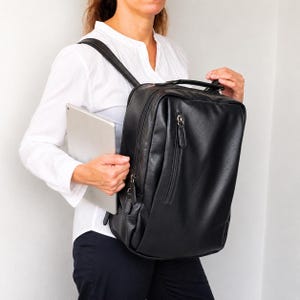 May include: A black rectangular backpack with a smooth finish, featuring a vertical zippered front pocket, a top handle, and adjustable shoulder straps. The backpack is shown alongside a silver tablet, highlighting its functional design for carrying electronics.