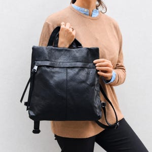 Può includere: Uno zaino nero di forma quadrata con finitura testurizzata, dotato di una patta frontale orizzontale, una cucitura centrale verticale e sottili cinghie laterali con dettagli metallici. La borsa include manici superiori e spallacci regolabili.