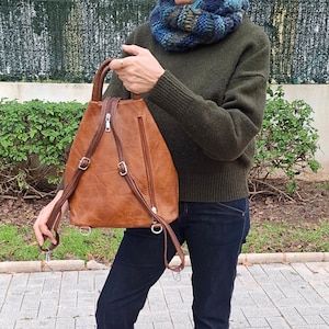 May include: A brown leather backpack with adjustable straps and a top handle. The backpack has a zippered main compartment and a front zippered pocket. The hardware is silver-colored. The bag is worn over the shoulders.