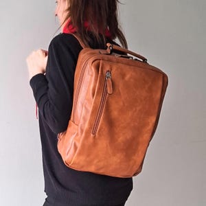 Pode incluir: Uma mochila retangular de couro castanho-claro com um bolso frontal vertical com fecho, uma alça superior e alças de ombro ajustáveis. A bolsa tem um acabamento mate liso com costuras visíveis nas bordas e fechos de metal.