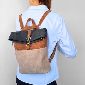 May include: A roll-top backpack featuring a color-blocked design with a dark charcoal gray top section, a medium brown middle panel, and a light beige bottom pocket. The bag includes brown shoulder straps, a top handle, and a brass-toned metal clasp.
