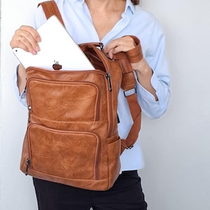 Puede incluir: Una mochila de color marrón claro con acabado texturizado, que cuenta con dos bolsillos frontales con cremallera, un compartimento principal con una tableta plateada en su interior y correas ajustables. La bolsa tiene cremalleras de metal oscuro.