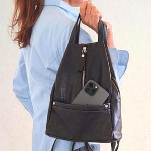 Könnte beinhalten: Ein schwarzer Rucksack mit einem vertikalen Reißverschlussfach auf der Vorderseite und einer offenen Außentasche. Die Tasche verfügt über zwei Tragegriffe und ein Smartphone in der Vordertasche, mit glatter, dunkler Oberfläche und silberfarbenen Beschlägen.