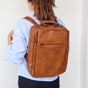 Puede incluir: Una mochila rectangular de color marrón claro con un acabado liso y ligeramente texturizado. La bolsa cuenta con un asa superior, correas ajustables para los hombros y un bolsillo frontal horizontal con cremallera. El compartimento principal tiene cierre de cremallera.