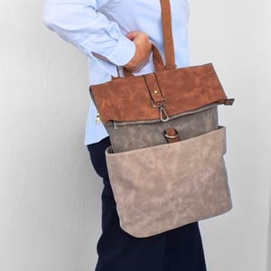 Op de afbeelding: Een meerkleurige rugzak met een bruine omslagflap met metalen sluiting, een donkergrijs middengedeelte en een lichtbeige ondervak. De tas is voorzien van bruine schouderbanden en een goudkleurige ritstrekker aan het zijvak.