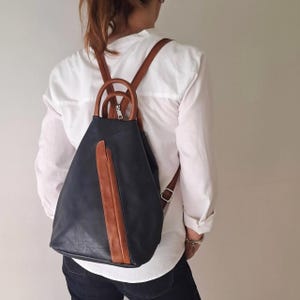 May include: A dark navy blue backpack with a vertical tan brown accent stripe down the centre. The bag features two rounded top handles and adjustable tan brown shoulder straps with silver metal buckles, creating a structured triangular shape.