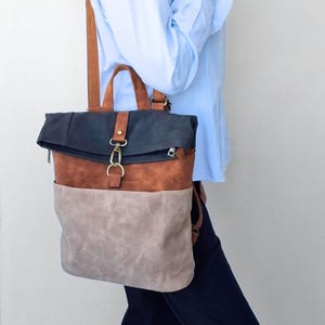 Puede incluir: Una mochila multicolor con cierre superior enrollable azul oscuro, una sección central marrón y un bolsillo inferior gris claro. La bolsa incluye correas marrones para los hombros, un asa superior y un cierre de metal dorado en la solapa.