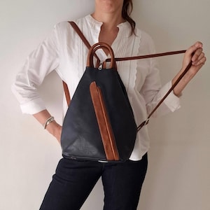 May include: A navy blue backpack with brown leather detailing and straps. The backpack features a rounded top with a brown handle and a vertical brown leather stripe. Adjustable straps are included.