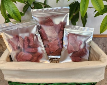 May include: Three silver, resealable bags filled with reddish-brown, dried food items are arranged in a beige basket with a green rim. The bags are of varying sizes, and the basket sits on a light-colored surface, with green leaves in the background.